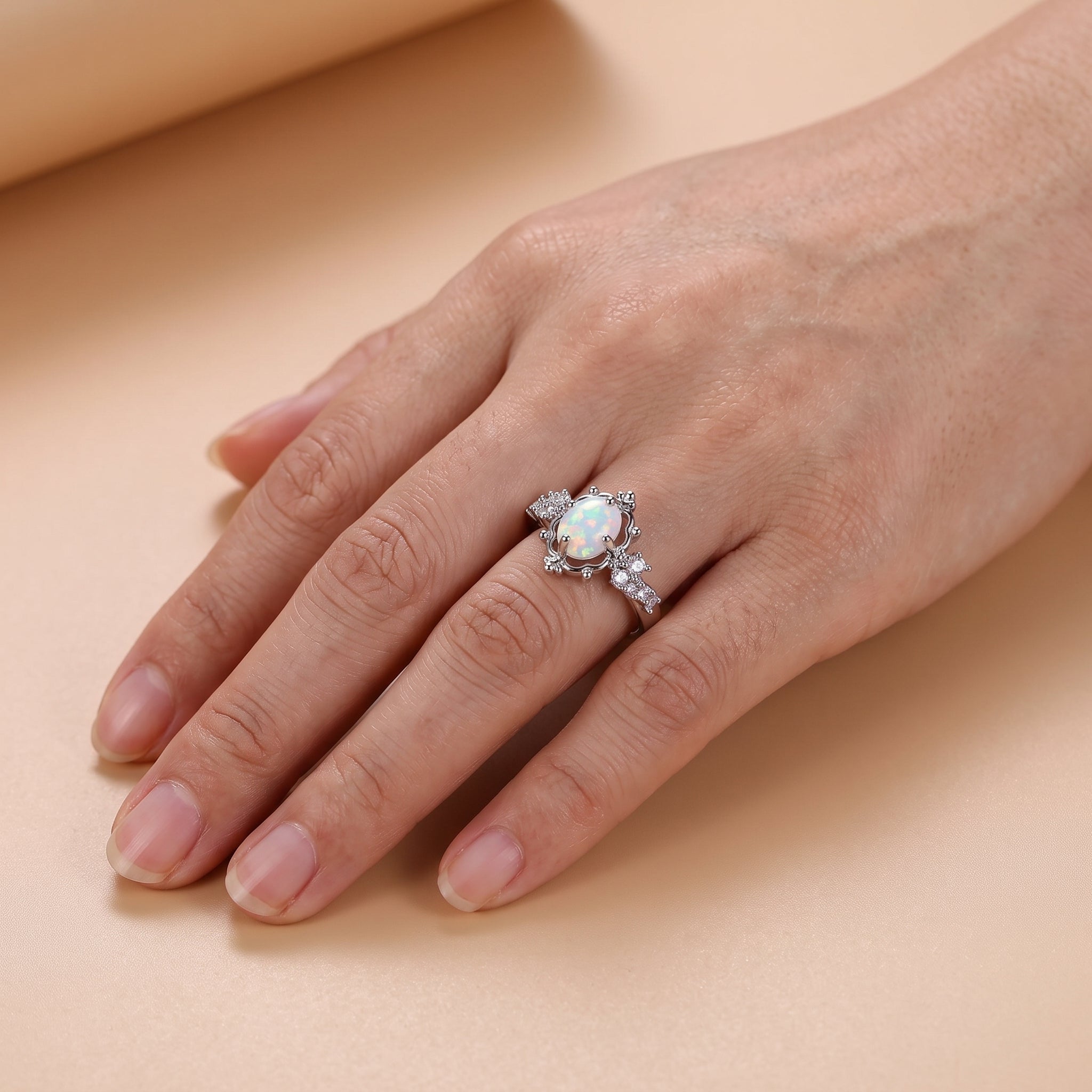 lifestyle: A new photorealistic photograph composed from scratch. The scene features a seamless matte warm beige studio surface that gently transitions into a softly graduated warm beige background. An adult hand, with clean, well-groomed nails in a natural finish, is positioned diagonally across the frame with the back of the hand facing the camera, fingers naturally and gently separated. The product is worn naturally on the ring finger, at a realistic scale, with correct physical contact to the skin, and…